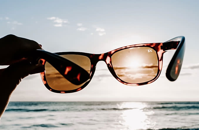 Hand holding sunglasses in front of sun at beach